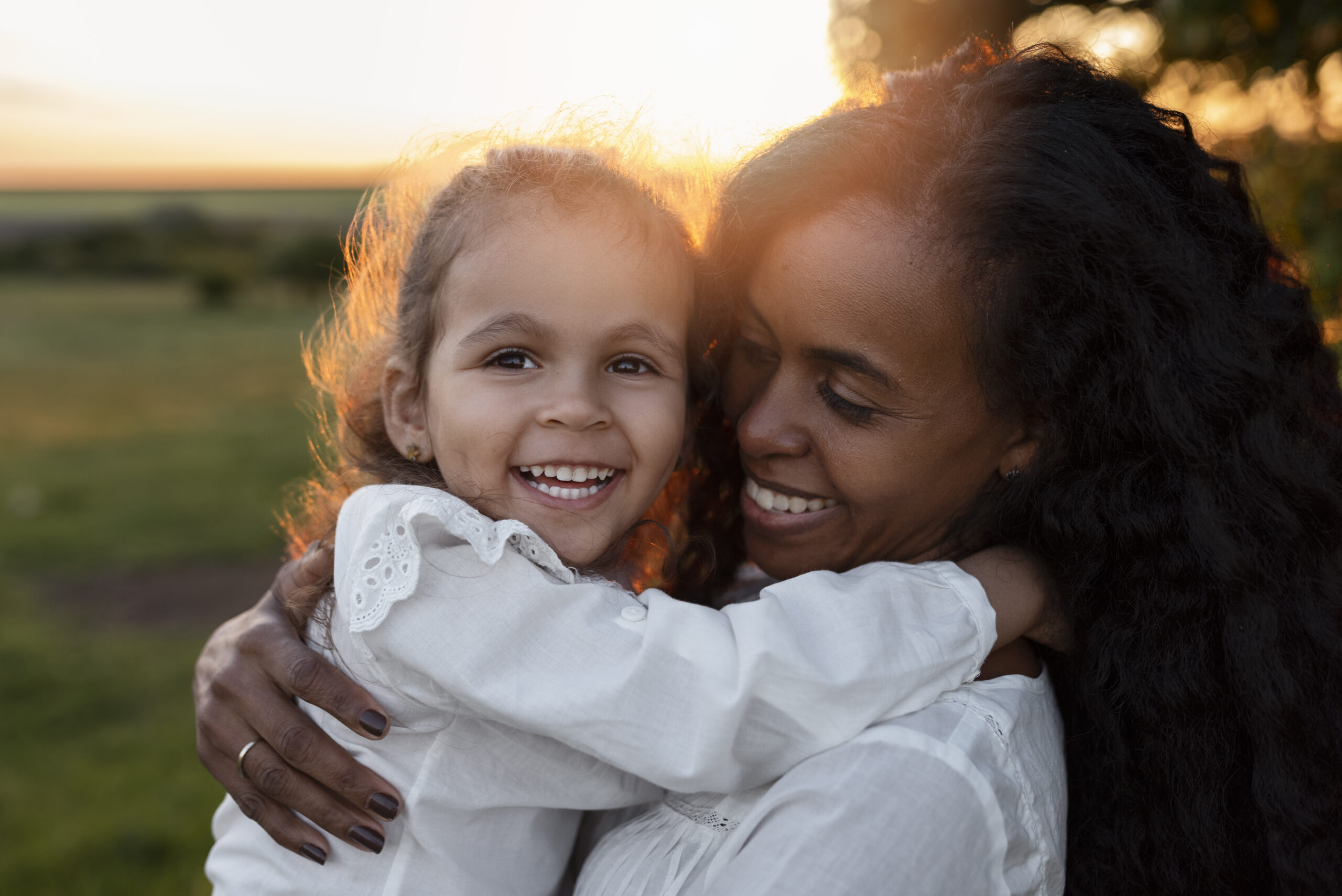 Mãe abraça a filha pequena ao pôr do sol em campo aberto — momento de afeto familiar