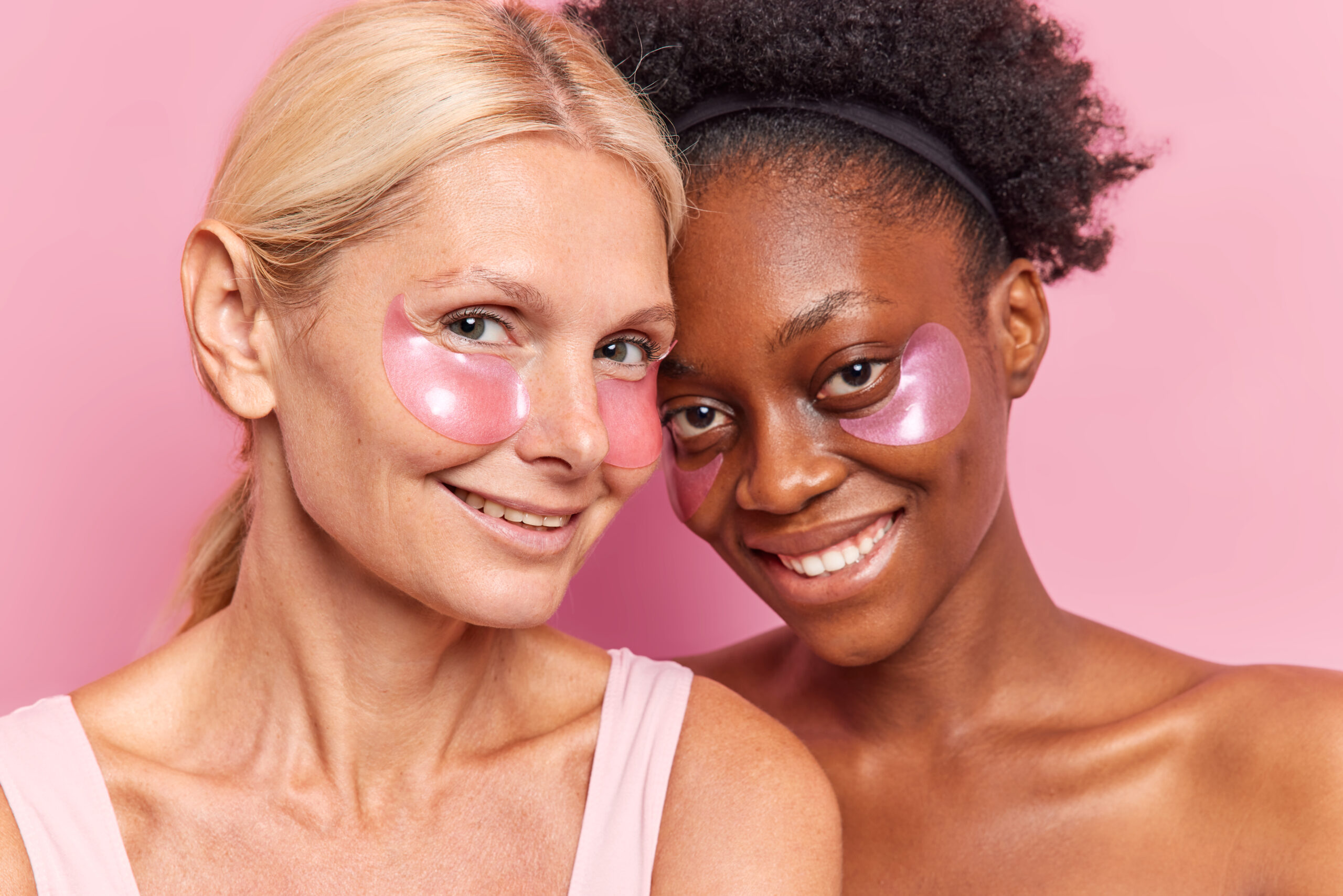 Duas mulheres de idades diferentes usam patches sob os olhos sorrindo em fundo rosa