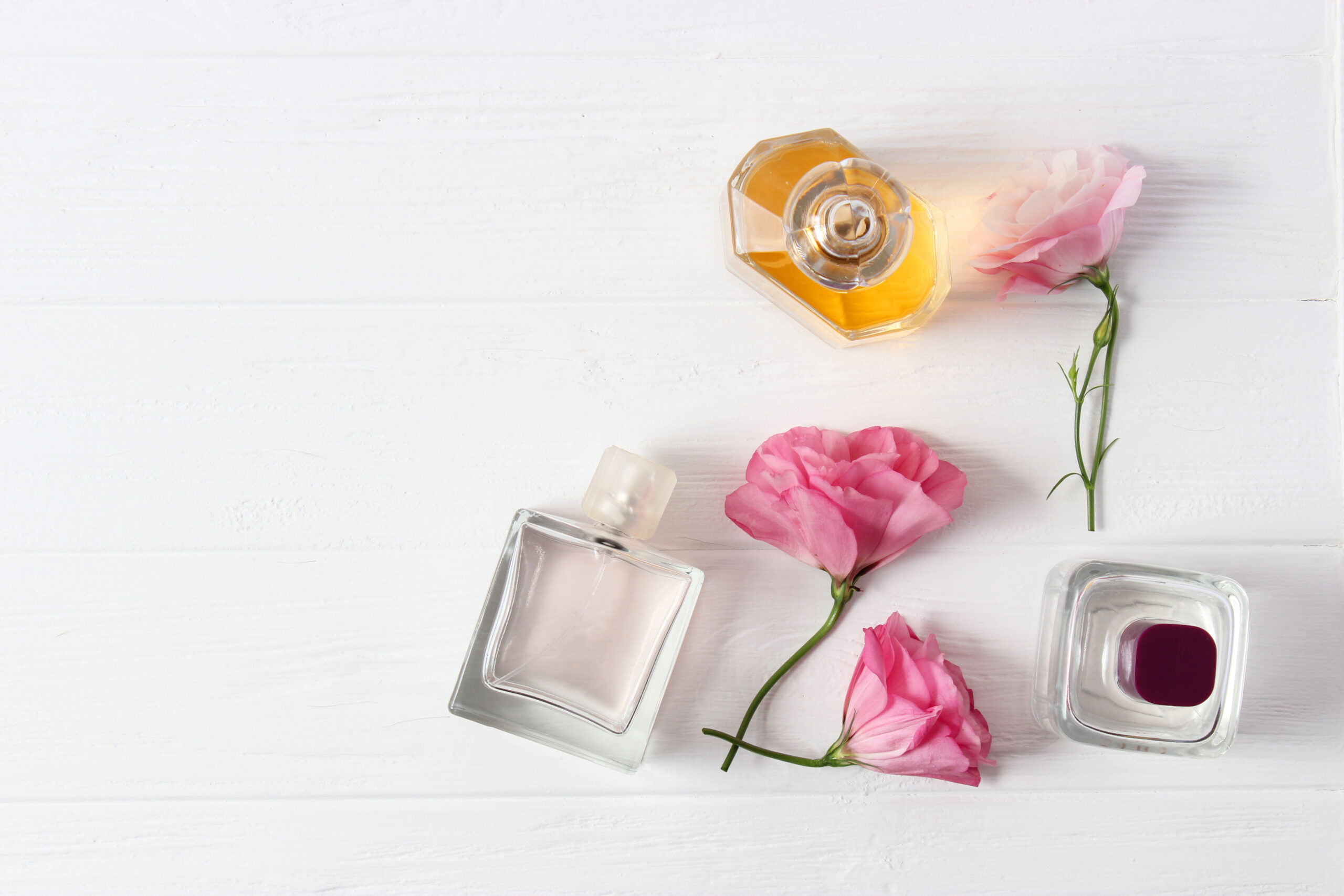 Três frascos de perfume em formatos e tons diferentes com rosas cor-de-rosa sobre fundo branco de madeira