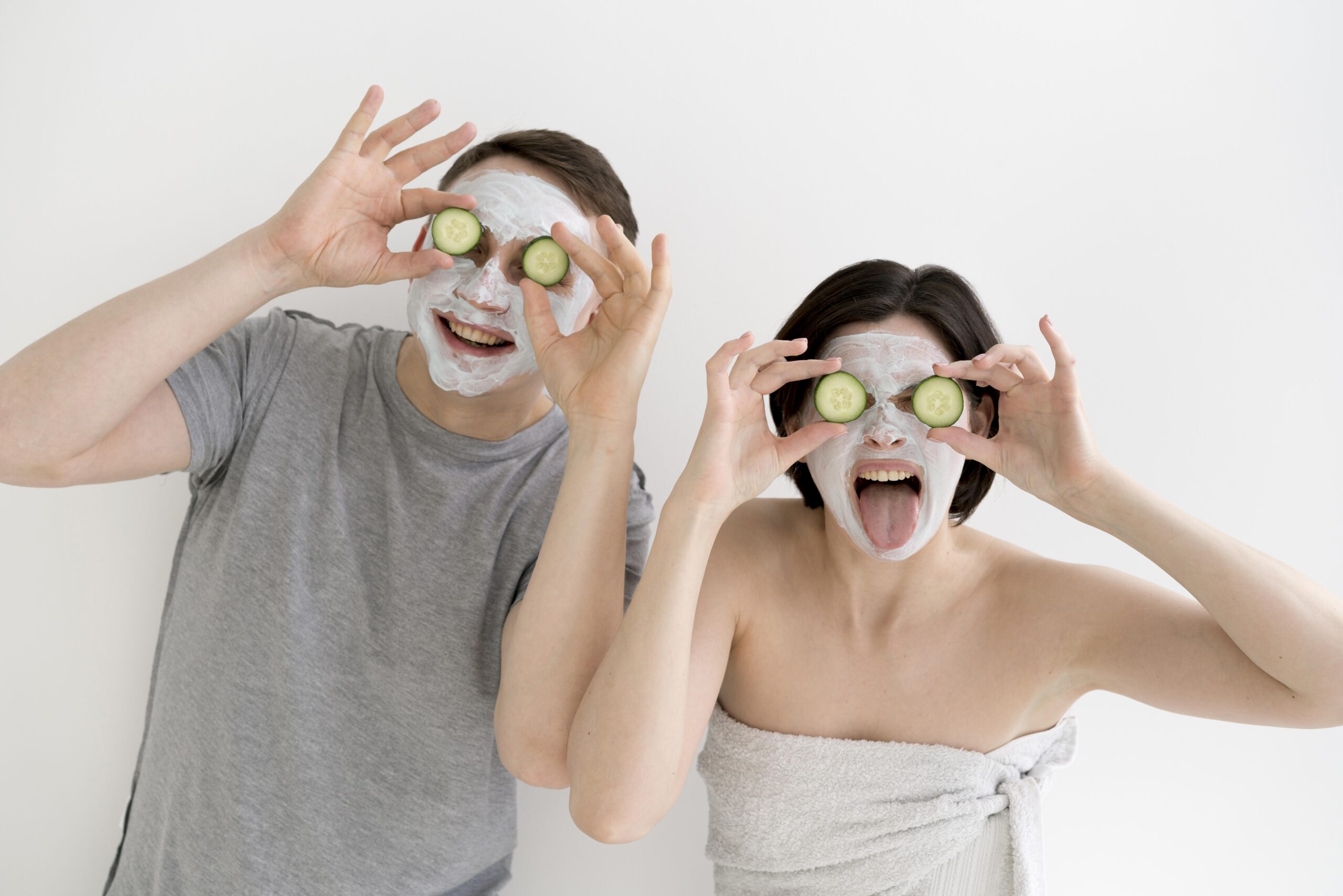 Casal sorri segurando fatias de pepino sobre os olhos com máscara facial branca no rosto
